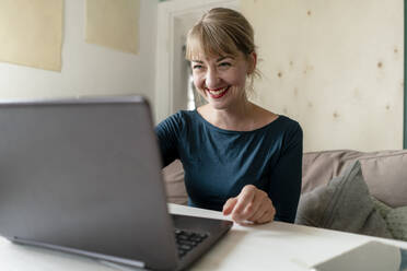 Porträt einer lachenden Frau, die zu Hause am Tisch sitzt und einen Laptop benutzt - KNSF06849