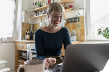 Porträt einer glücklichen Frau, die in der Küche sitzt und einen Laptop benutzt - KNSF06850