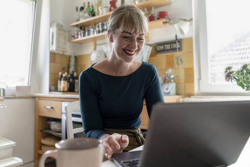 Portrait of happy woman sitting in the kitchen using laptop - KNSF06850