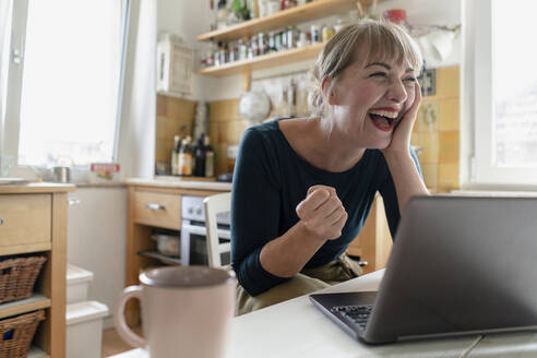 Portrait of woman sitting in the kitchen with laptop crying for joy - KNSF06854