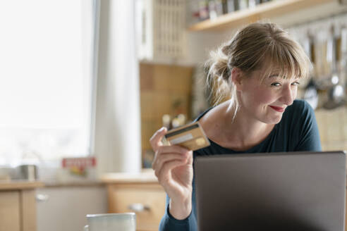 Lächelnde Frau in der Küche mit Kreditkarte und Laptop - KNSF06857
