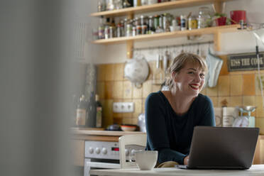 Portrait of smiling woman sitting in the kitchen using laptop - KNSF06858