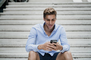 Junger Mann sitzt auf einer Treppe und benutzt sein Smartphone - JMHMF00023
