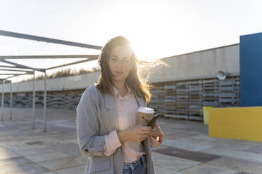 Porträt einer jungen Frau mit Coffee to go und Mobiltelefon im Gegenlicht - AFVF04082