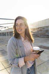Porträt einer lächelnden jungen Frau mit Kaffee zum Mitnehmen und Mobiltelefon, die in die Ferne schaut - AFVF04083