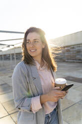 Portrait of smiling young woman with coffee to go and mobile phone looking at distance - AFVF04083