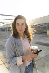 Portrait of young woman with coffee to go and mobile phone at backlight - AFVF04084