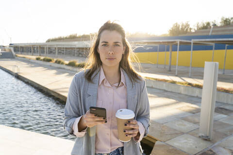 Porträt einer ernsten jungen Frau mit Mobiltelefon und Kaffee zum Mitnehmen, lizenzfreies Stockfoto