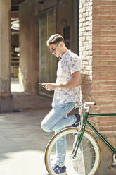 Man with bicycle leaning against a wall using his smartphone - JNDF00144