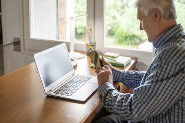 Älterer Mann sitzt zu Hause vor einem Laptop und benutzt ein Smartphone - AFVF04107