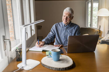 Älterer Mann sitzt zu Hause vor einem Laptop und macht sich Notizen in einem Notizbuch - AFVF04110