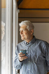 Lächelnder älterer Mann, der durch ein Fenster auf sein Haus blickt - AFVF04116