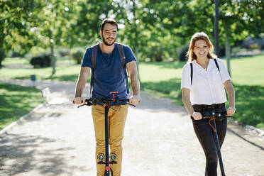 Portrait of content couple riding electric scooters in a city park - JSMF01318