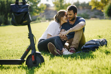 Happy couple with electric scooter relaxing on a meadow in a park - JSMF01326