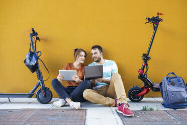 Portrait of couple with backpacks and electric scooters using laptop and digital tablet - JSMF01343