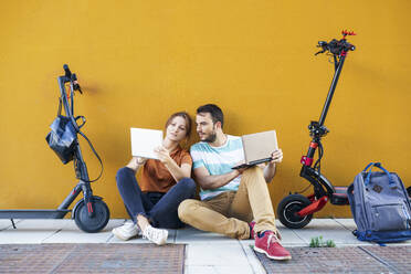 Portrait of couple with backpacks and electric scooters using laptop and digital tablet - JSMF01344