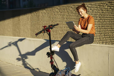 Smiling woman with electric scooter sitting on a wall at sunlight, using digital tablet - JSMF01352