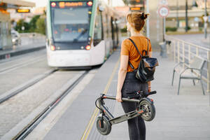 Back view of woman with backpack and folded electric scooter waiting at tram stop - JSMF01361