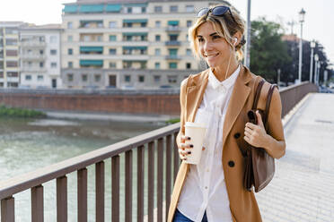 Smiling woman with earphones and coffee to go walking through city - GIOF07311
