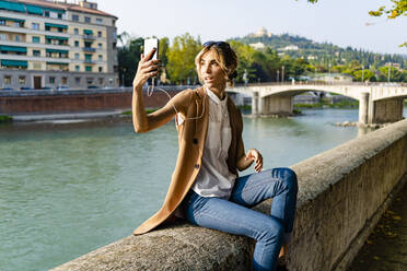 Frau mit Kopfhörern sitzt am Flussufer und macht ein Selfie - GIOF07314