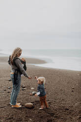 Mother with baby in carrier playing with toddler on beach - ISF22327