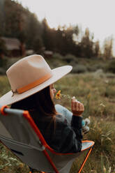 Young woman in stetson smelling wildflower on deck chair in rural valley, rear view, Mineral King, California, USA - ISF22362