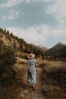 Junge Frau mit Stetson und Maxikleid, die auf einem Feldweg in einem ländlichen Tal spazieren geht, Rückansicht, Mineral King, Kalifornien, USA - ISF22384