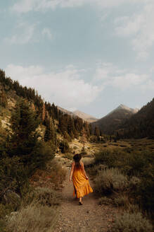 Frau in orangefarbenem Maxikleid schlendert über einen Feldweg in einem ländlichen Tal, Rückansicht, Mineral King, Kalifornien, USA - ISF22385