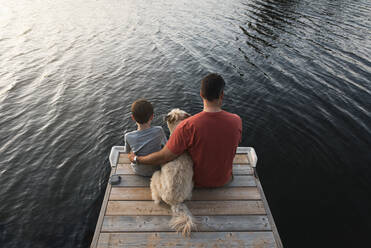Vater und Sohn sitzen mit ihrem Hund am Ende eines Stegs an einem See. - CAVF65699