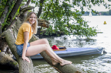 Lächelnde Frau sitzt auf einem Baumstamm am Seeufer - BFRF02101