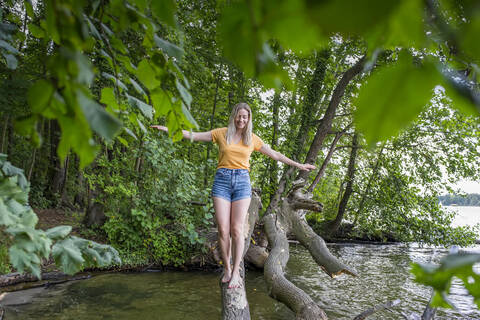 Lächelnde Frau balanciert auf einem Baumstamm am Seeufer, lizenzfreies Stockfoto