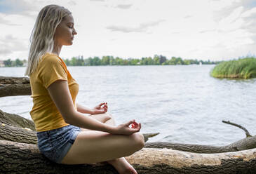 Junge Frau meditiert auf einem Baumstamm am Seeufer - BFRF02111