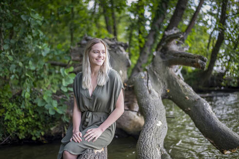 Lächelnde Frau sitzt auf einem Baumstamm am Seeufer - BFRF02132