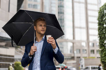 Junger Geschäftsmann mit Regenschirm trinkt einen Kaffee an einem regnerischen Tag in Bangkok - MAUF02981