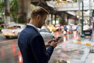 Young businessman using smartphone in Bangkok during a rainy day - MAUF03008