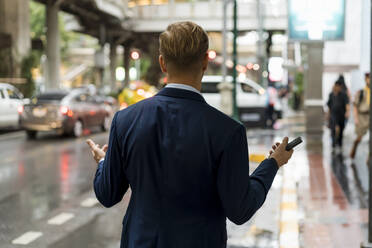 Young businessman holding smartphone in Bangkok during a rainy day - MAUF03009