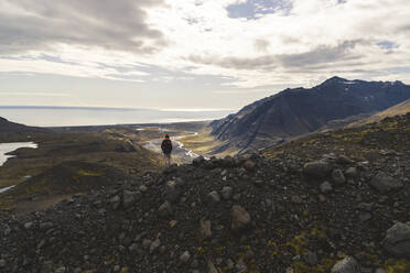 A hiker stands near the edge of a cliff at the base of Jöklasel Glacier, overlooking the valley towards the Southern Region of Iceland. - AAEF04596