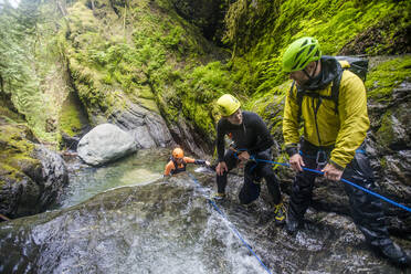 Drei abenteuerlustige Männer arbeiten als Team, um einen Freund in einem Wasserfall zu sichern. - CAVF65786