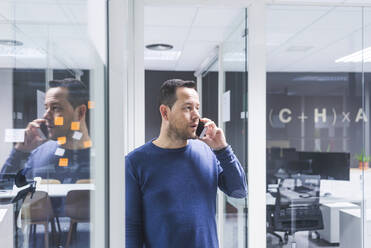 Side view of businessman talking on smart phone while standing against office wall - CAVF65852