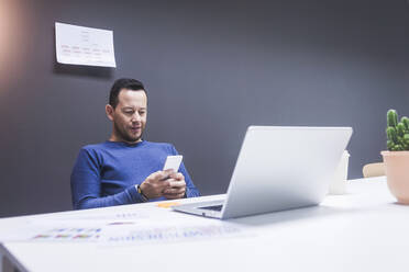 Junger Geschäftsmann liest sein Smartphone. Porträt eines Geschäftsmannes, der eine Nachricht mit seinem Smartphone im Büro liest. Ein Mann arbeitet an seinem Schreibtisch im Büro. - CAVF65855