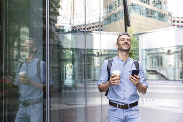 Smiling businessman holding cup of coffee and smartphone in the city, Berlin, Germany - WPEF02067