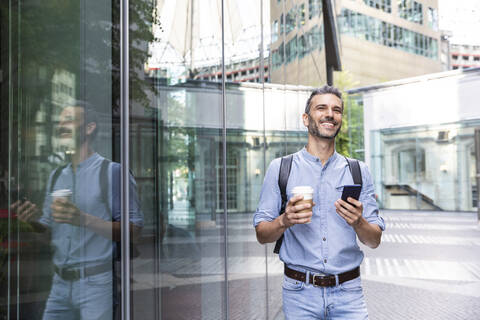 Lächelnder Geschäftsmann mit Tasse Kaffee und Smartphone in der Stadt, Berlin, Deutschland, lizenzfreies Stockfoto