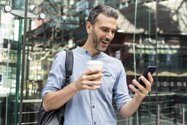 Glücklicher Geschäftsmann mit Blick auf das Smartphone in der Stadt, Berlin, Deutschland - WPEF02071