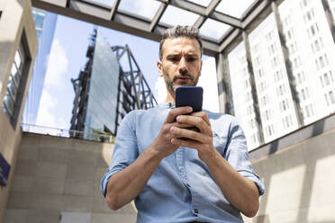 Seriöser Geschäftsmann mit Blick auf das Smartphone in der Stadt, Berlin, Deutschland - WPEF02072