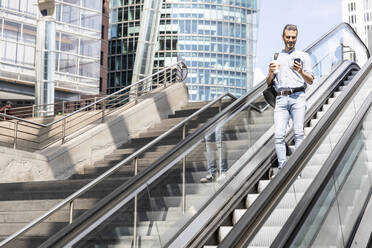 Lächelnder Geschäftsmann mit Blick auf das Smartphone auf einer Rolltreppe in der Stadt, Berlin, Deutschland - WPEF02076