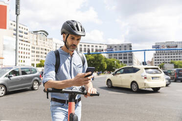 Businessman looking at the smartphone commuting on electric scooter in the city, Berlin, Germany - WPEF02081