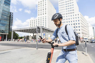 Businessman looking at the smartphone commuting on electric scooter in the city, Berlin, Germany - WPEF02082