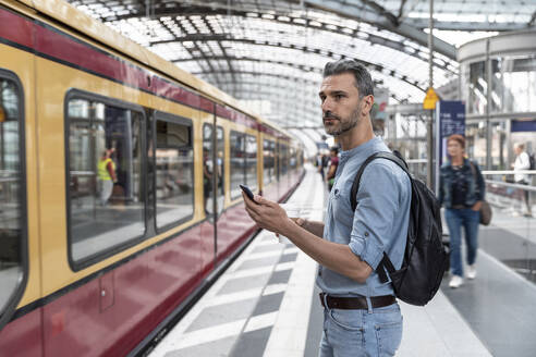 Geschäftsmann mit Smartphone auf dem Bahnhof bei der Einfahrt eines Zuges, Berlin, Deutschland - WPEF02095