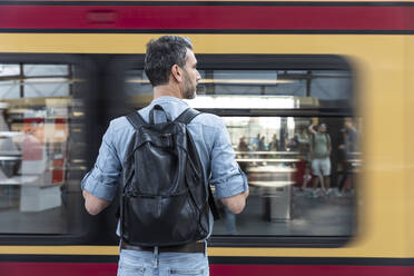 Rear view of man with backpack at the station platform while train coming in, Berlin, Germany - WPEF02096