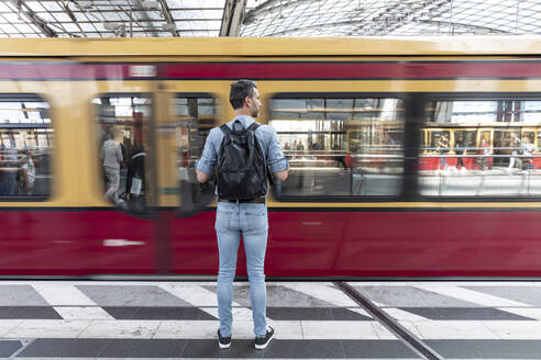 Rückansicht eines Mannes mit Rucksack auf dem Bahnsteig bei der Einfahrt eines Zuges, Berlin, Deutschland - WPEF02097